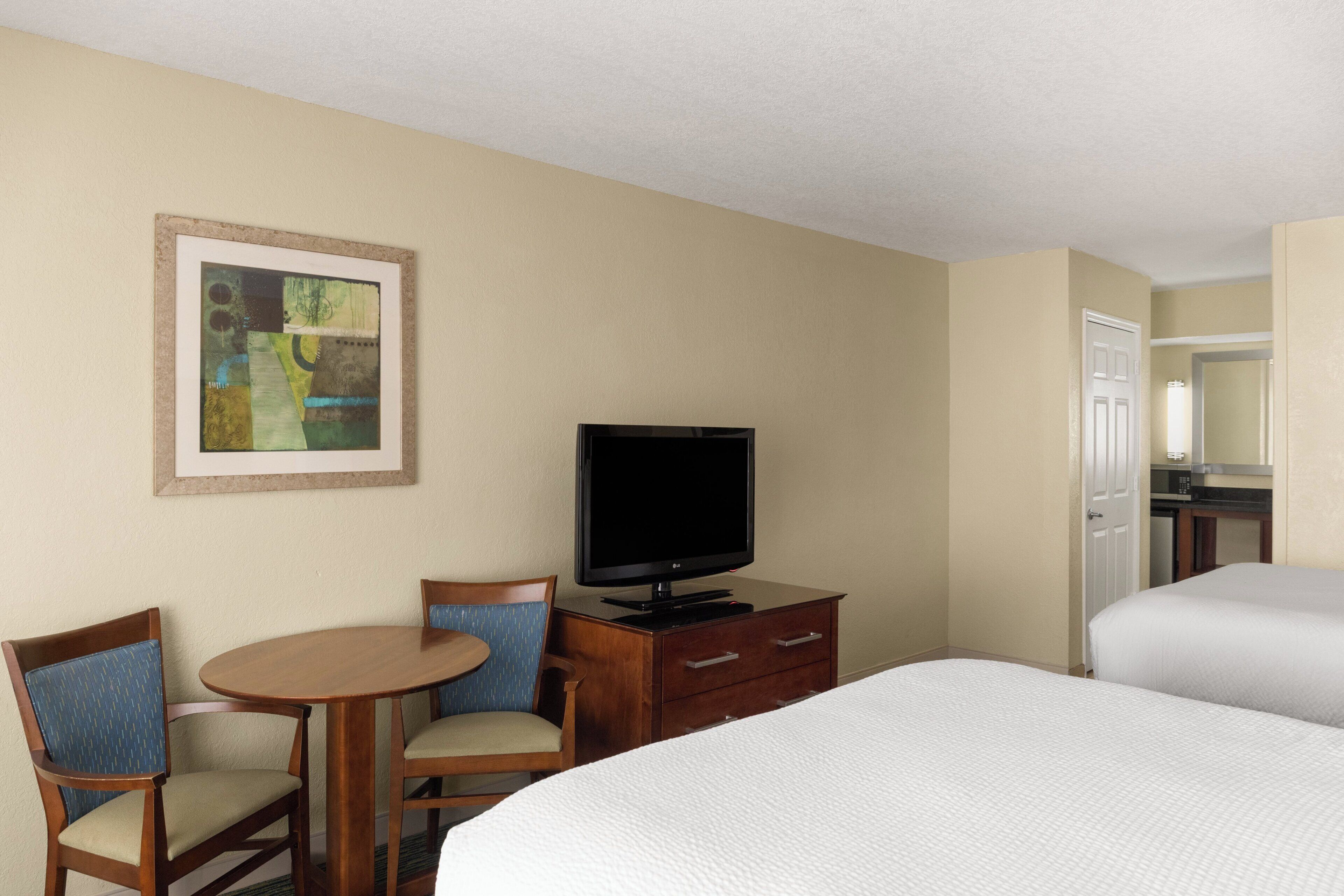 A hotel room with a bed, a television on a wooden dresser, a small table with two chairs, and a painting on the wall.