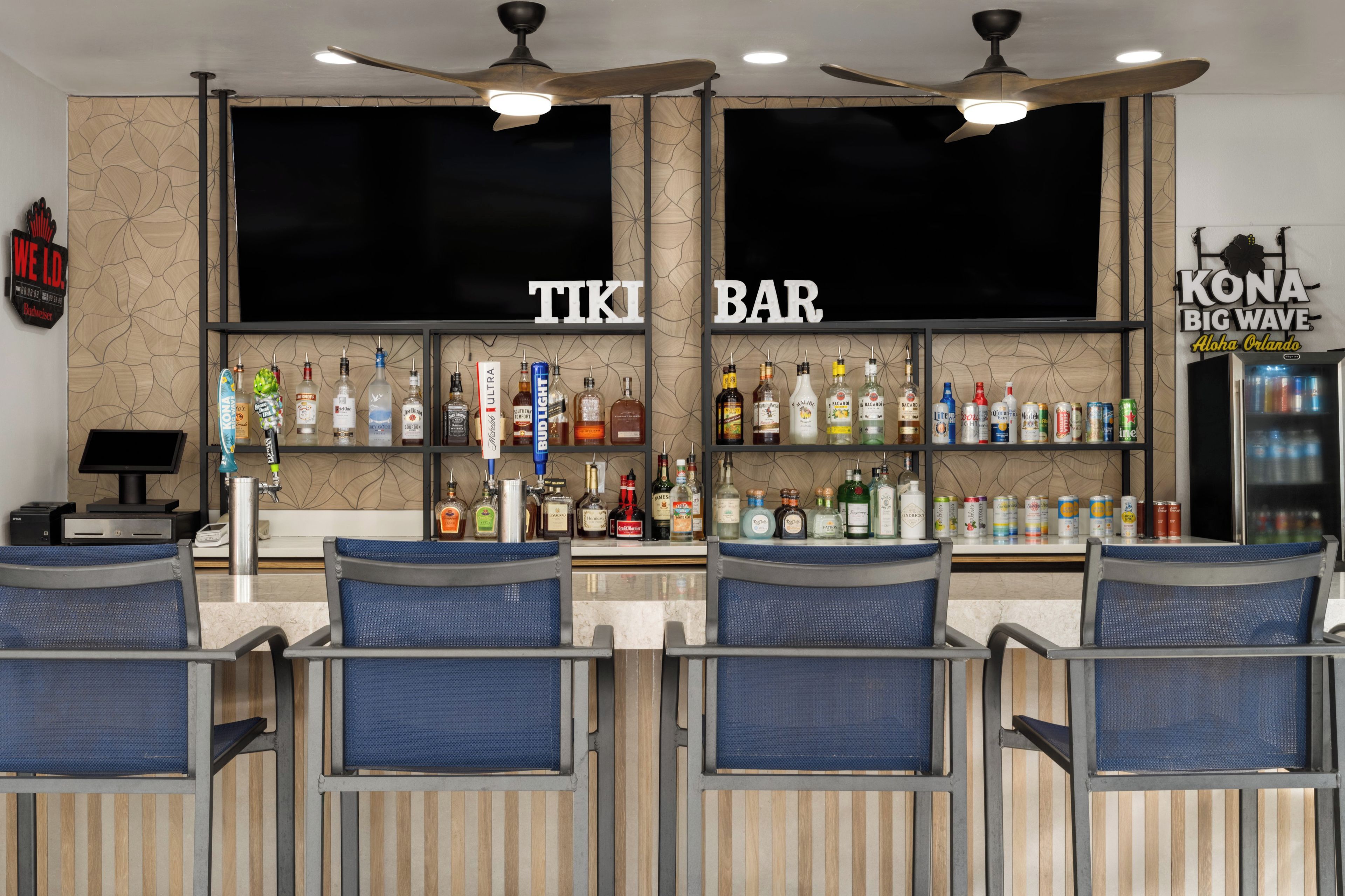 Een bar met twee grote schermen, diverse drankflessen en zitplaatsen met blauwe stoelen.