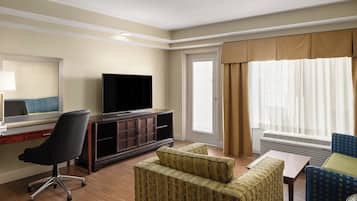 Quarto de hotel com televisão de tela plana, escrivaninha, cadeira, sofá e janela com cortinas.