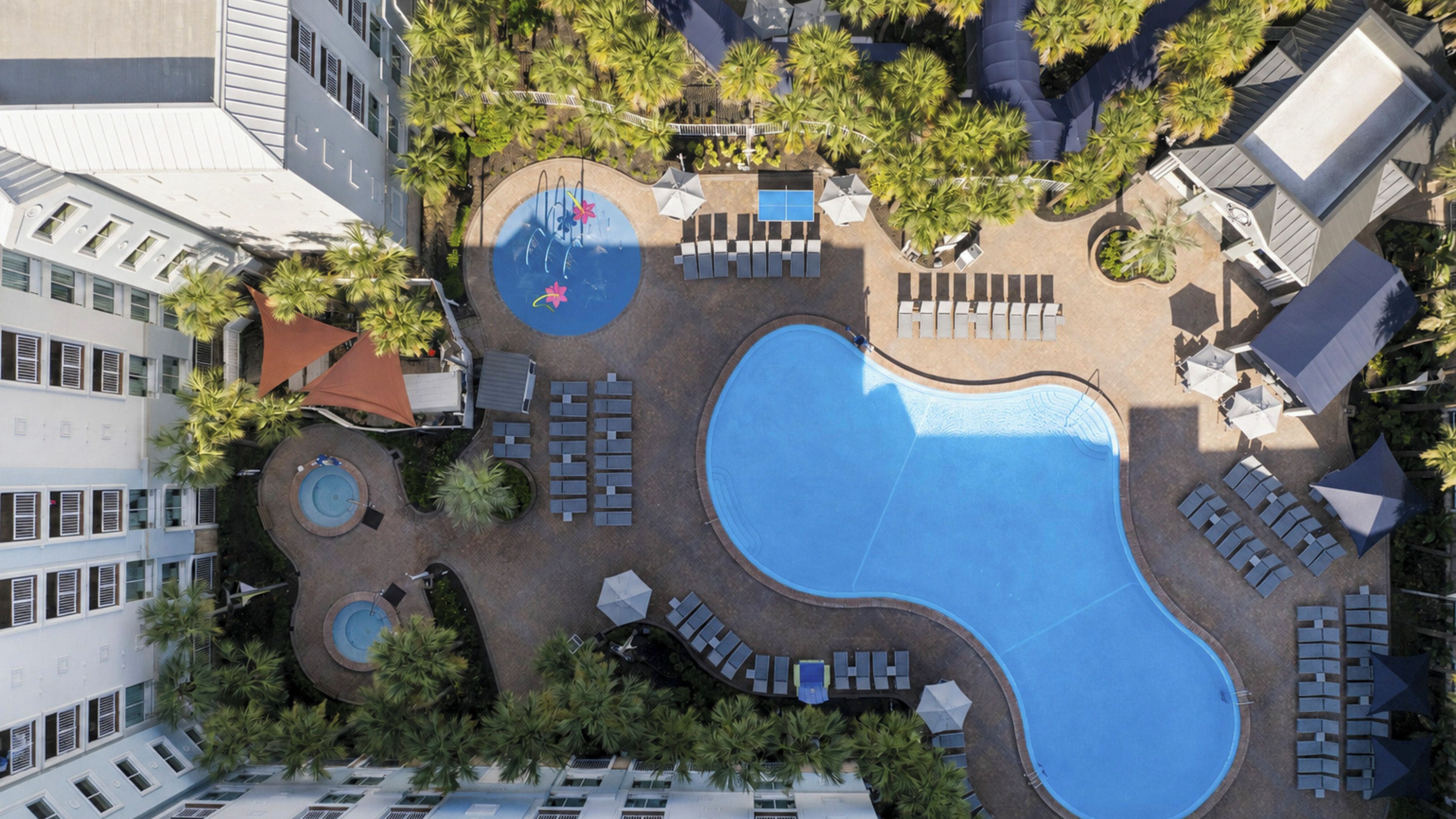 An aerial view of a hotel with a large swimming pool, sun loungers, and palm trees.