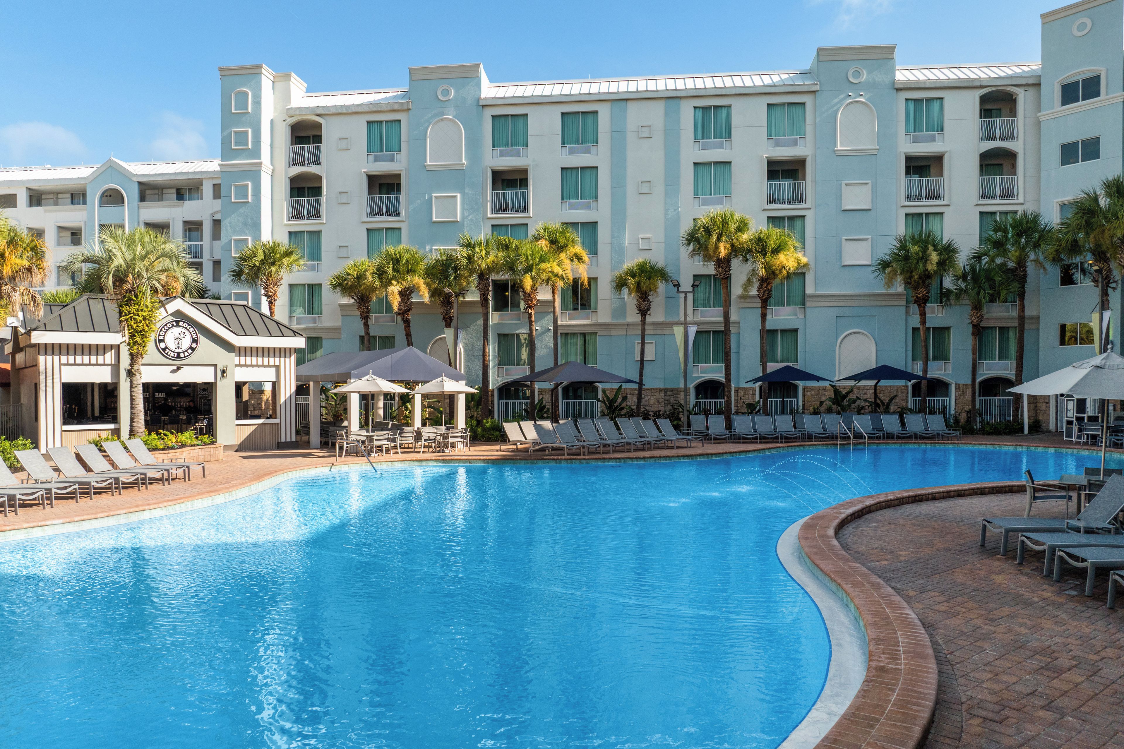 A hotel pool area with lounge chairs and umbrellas, surrounded by palm trees and a multi-story building with balconies.