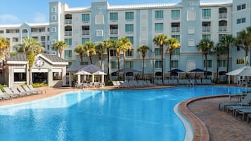 A hotel pool area with lounge chairs and umbrellas, surrounded by palm trees and a multi-story building with balconies.