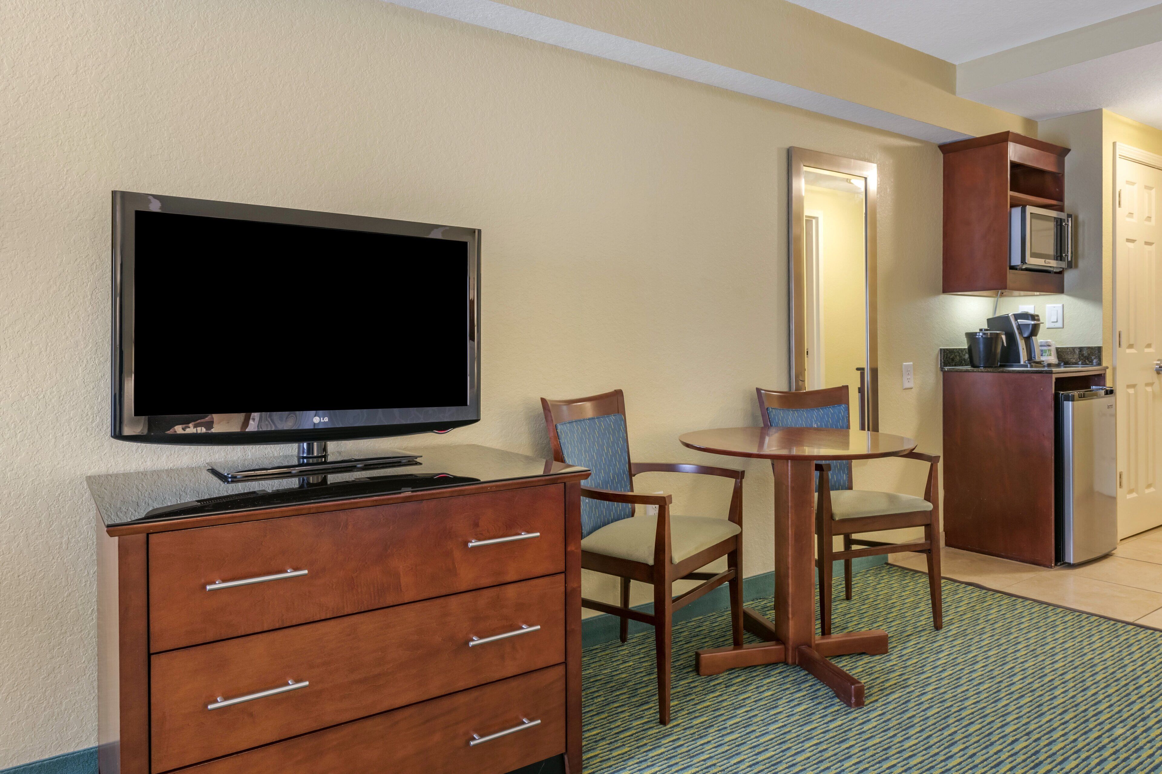 Ein Hotelzimmer mit Flachbildfernseher, einem Holzkasten, zwei Stühlen, einem kleinen Tisch, einer Mikrowelle und einem Kühlschrank.