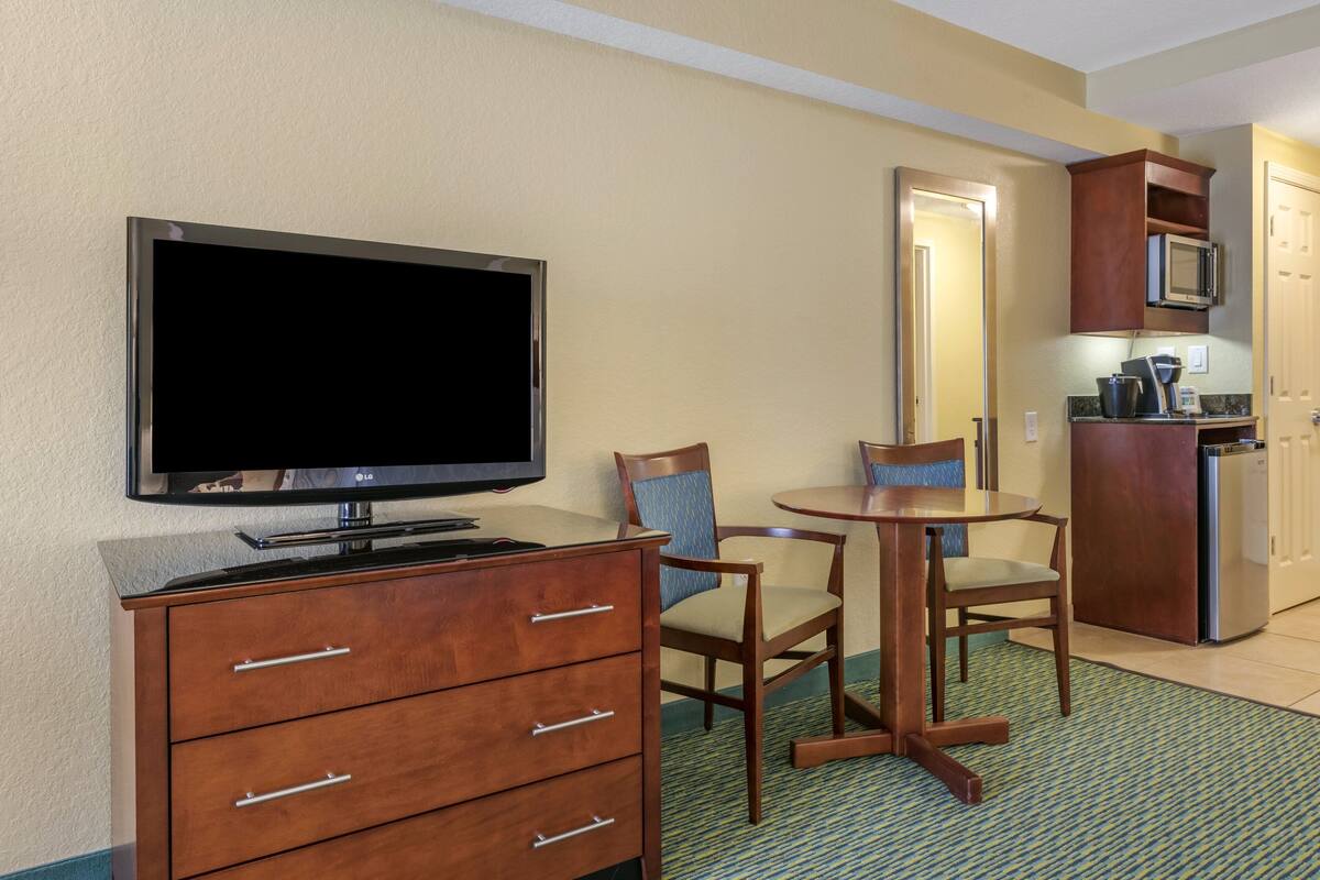 A hotel room with a flat-screen TV, wooden dresser, two chairs, a small table, a microwave, and a refrigerator.