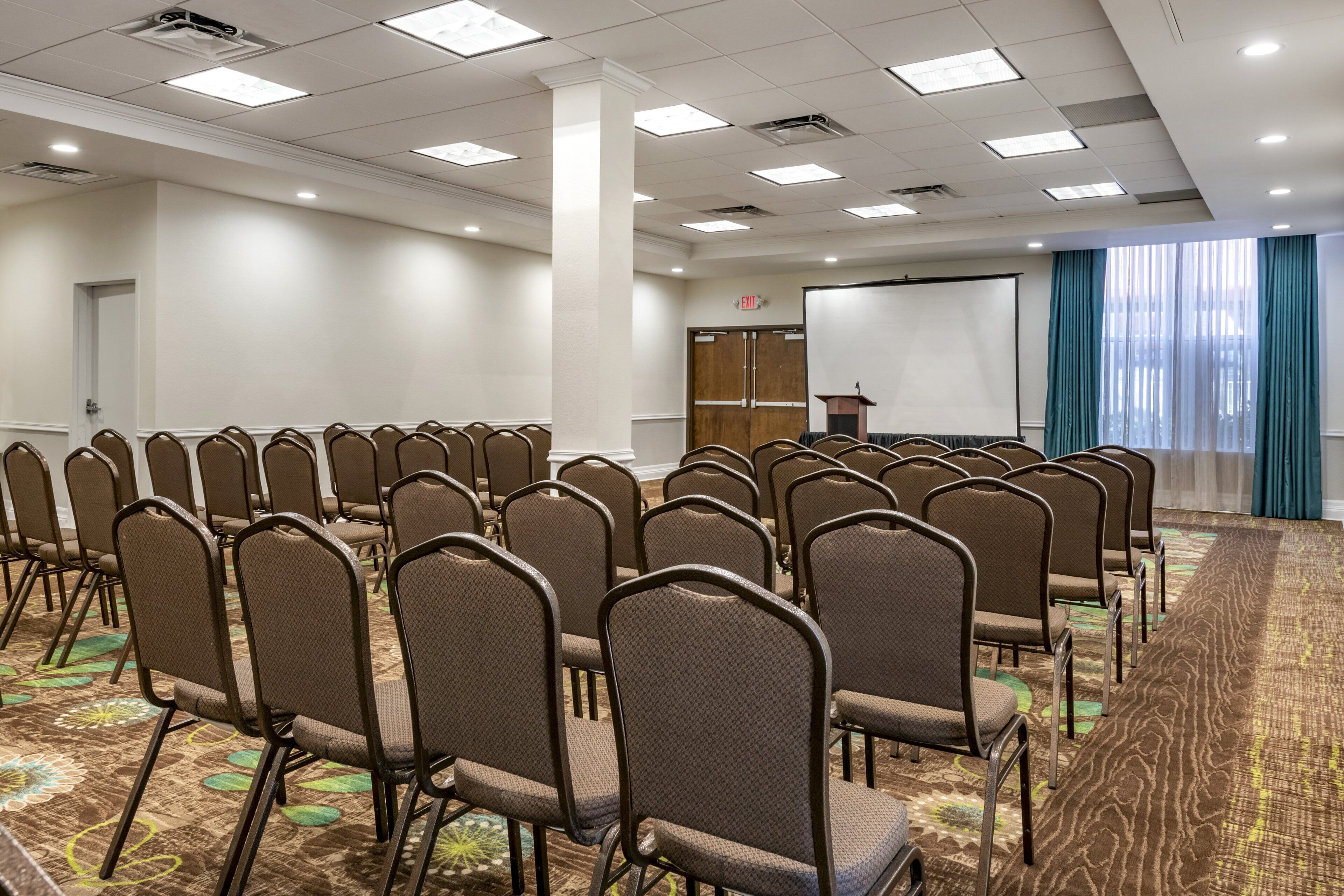 A conference room with rows of chairs, a podium, a projection screen, and a carpeted floor.