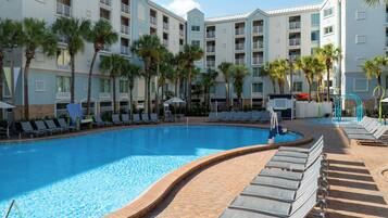 Una piscina con sillas de descanso y palmeras, rodeada por un edificio hotelero de varios pisos.