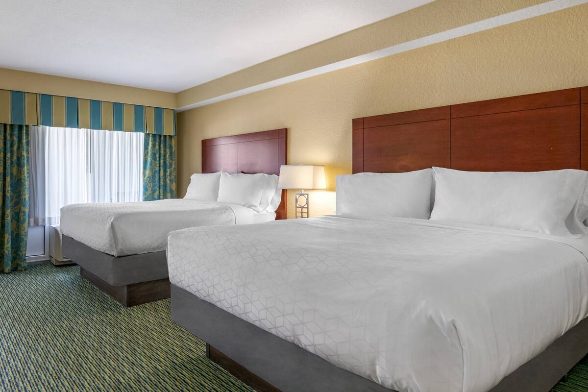 A hotel room with two beds, a wooden headboard, a lamp, and a window with curtains.