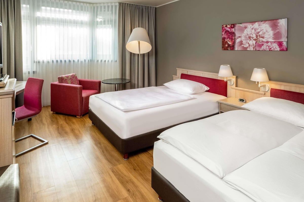 A hotel room with two beds, a desk, a chair, a small table, and a lamp.