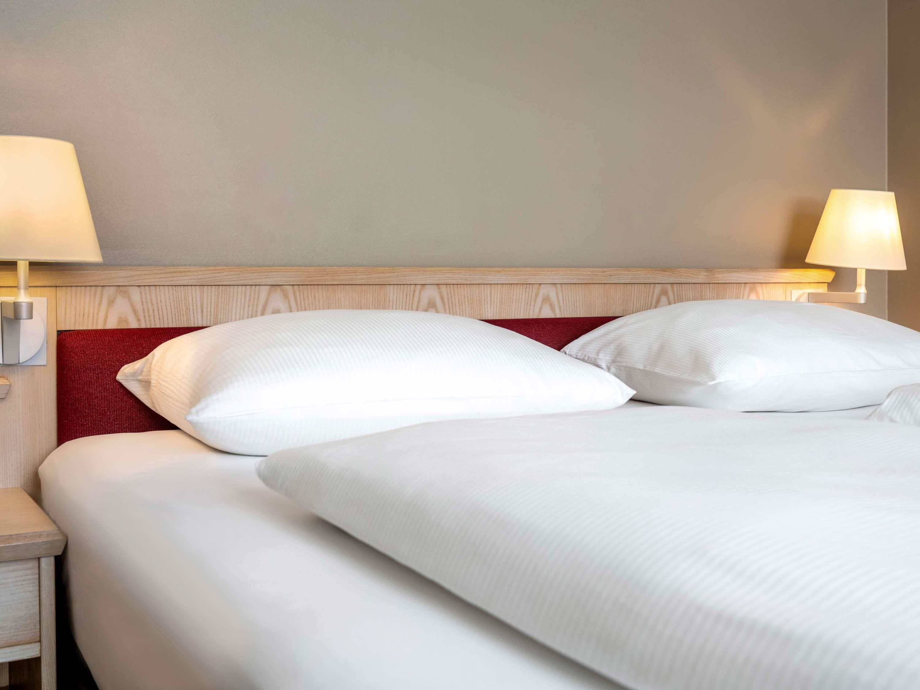 Cama bem arrumada com lençóis brancos e uma almofada vermelha de destaque, acompanhada por duas luminárias de cabeceira em uma mesinha de madeira.