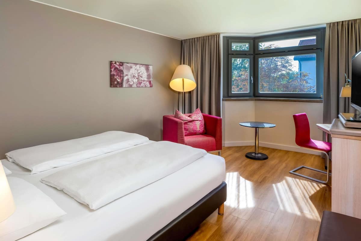 A hotel room with a bed, a desk, a chair, a lamp, a window, and a painting on the wall.