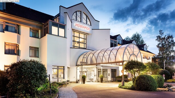 A modern hotel with a curved glass facade, a prominent entrance, and a well-maintained garden area. - Mövenpick Hotel München Airport (Hallbergmoos)