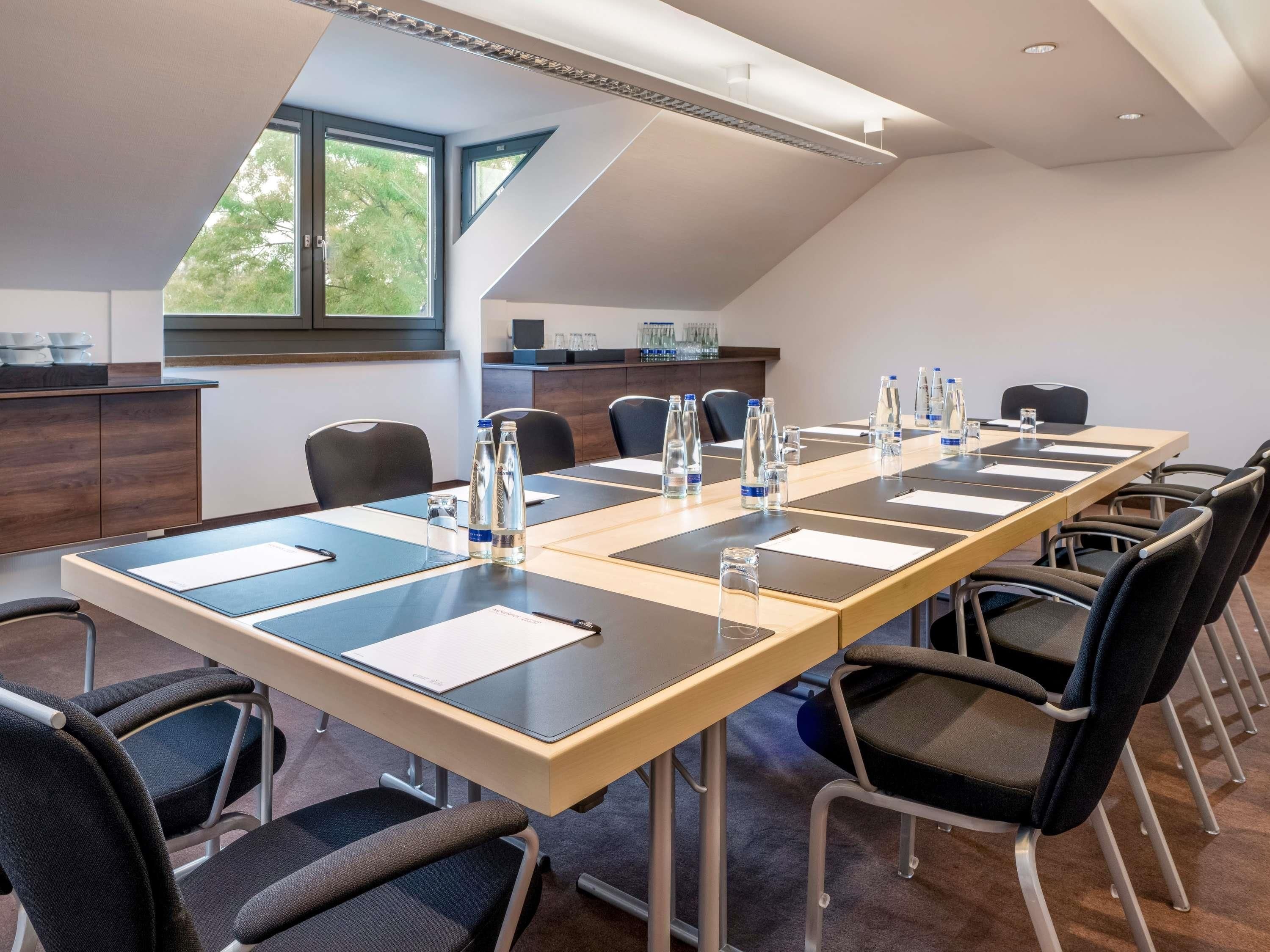 A conference room with a long table, chairs, water bottles, and a view of trees through a window.
