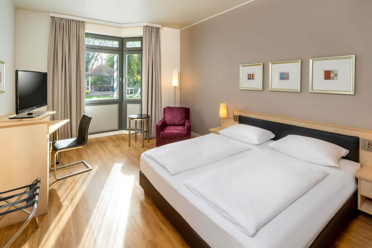 A hotel room with a large bed, a desk with a TV, a chair, a small table, and a window with curtains.
