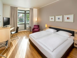 Una habitación de hotel con una cama grande, un escritorio con televisor, una silla, una mesita y una ventana con cortinas.