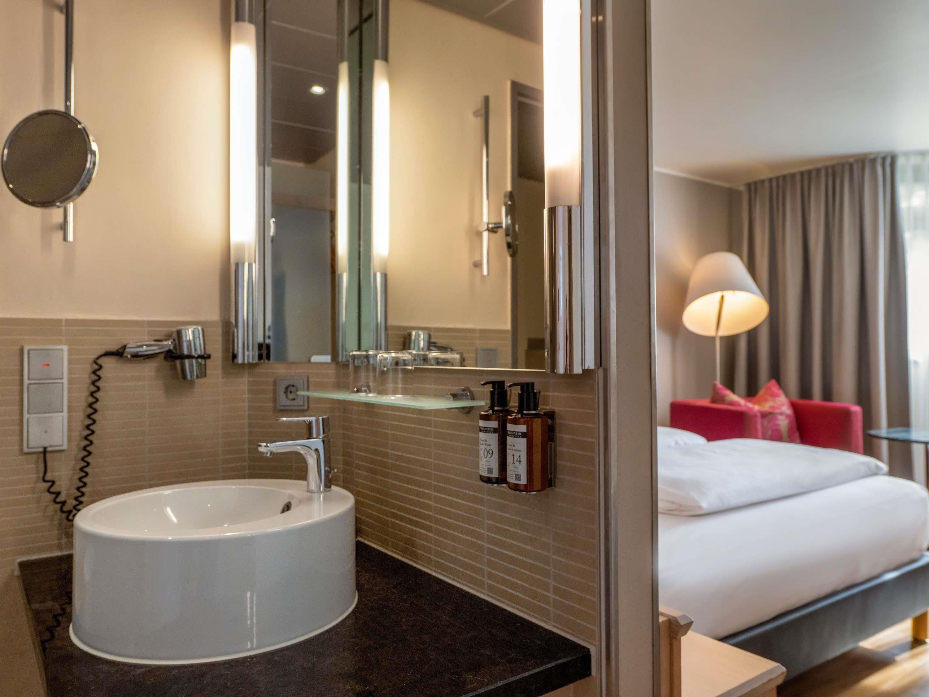 Un baño de hotel con un lavamanos blanco, un espejo y un secador de pelo. Se ve una cama con cabecera roja y una lámpara a un lado.