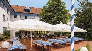 An outdoor dining area with tables covered by white umbrellas, arranged on a brick patio with a building in the background. - Mövenpick Hotel München Airport (Hallbergmoos)