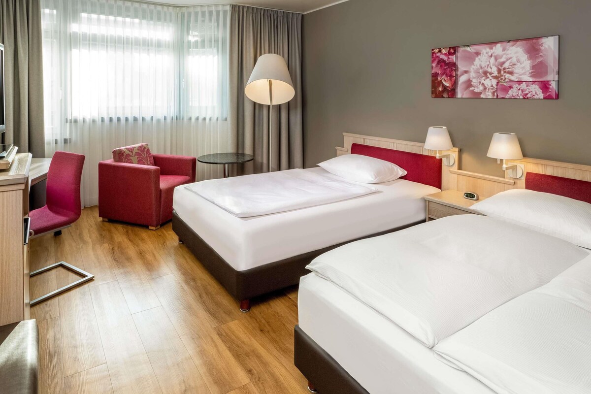 A hotel room with two beds, a desk, a chair, a small table, and a lamp.
