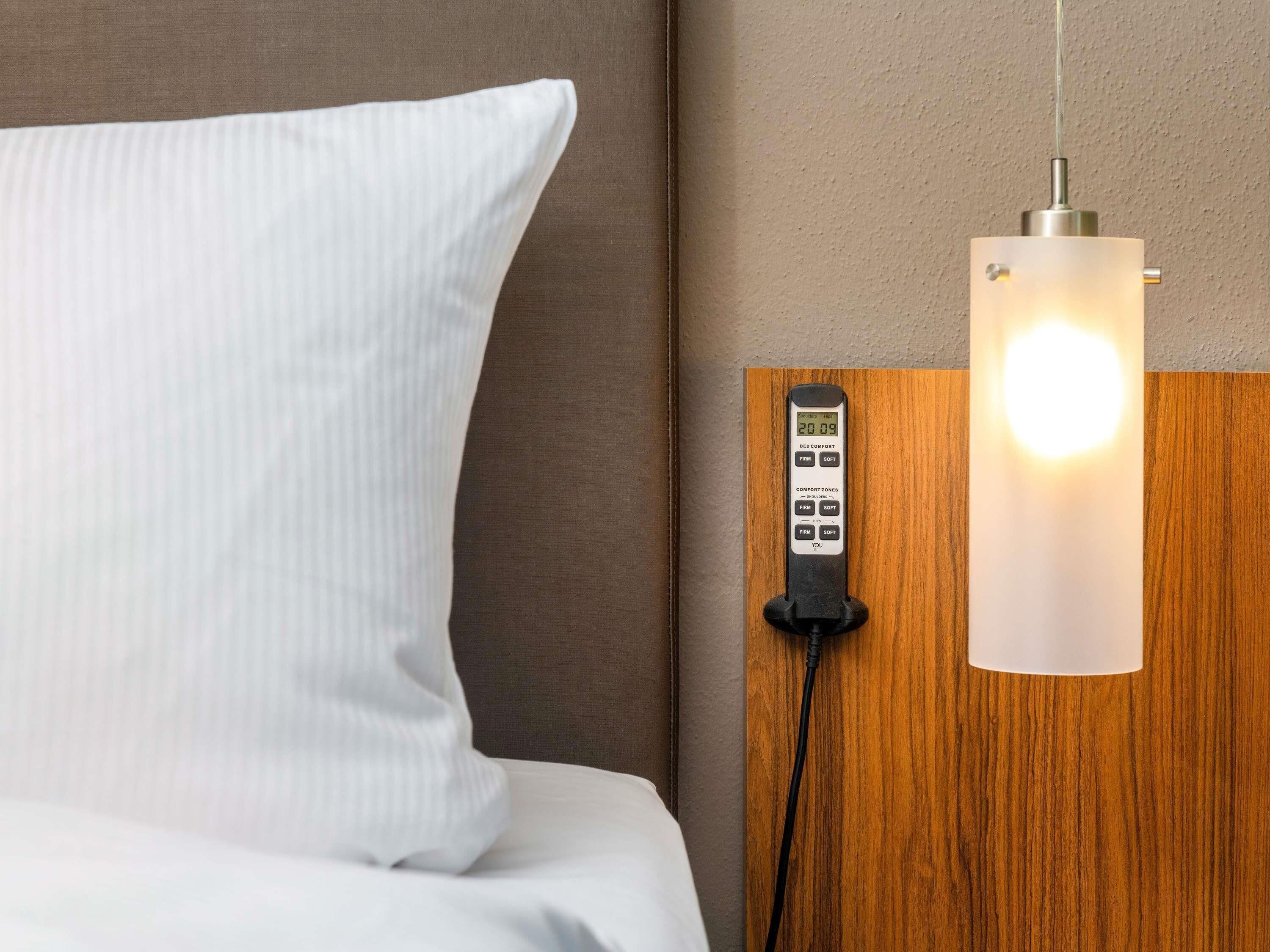A bed with a white pillow, a bedside control panel, and a wall-mounted light fixture.