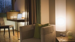 A modern room with a sofa, a side table with wine glasses, a lamp, and a laptop on a desk. - Mövenpick Hotel München Airport (Hallbergmoos)