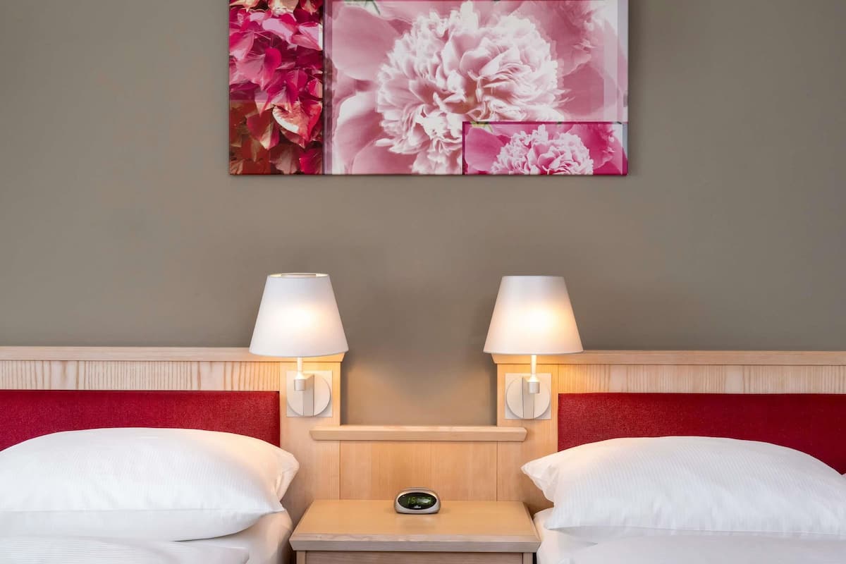 Quarto de hotel com duas camas, uma mesa de cabeceira com relógio digital e luminárias fixadas na parede.