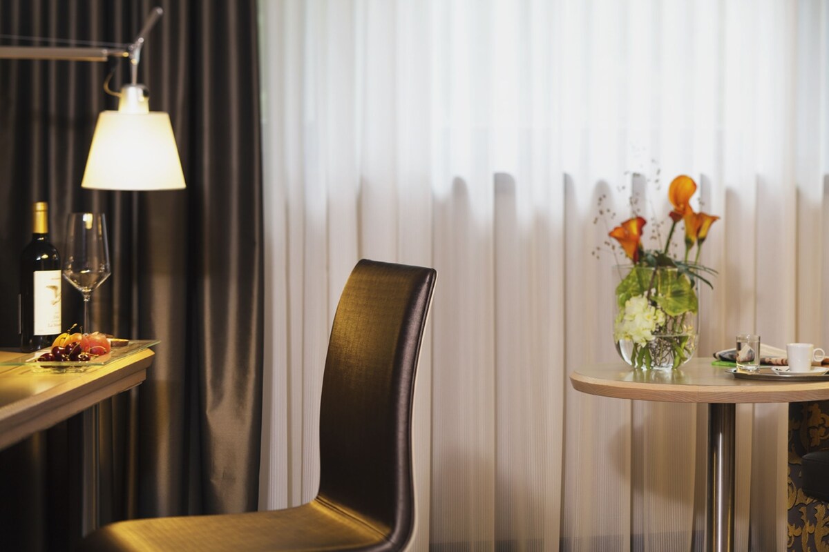 A hotel room with a dining table set with a bottle of wine, a glass, and a plate of fruit. There is a chair and a small round table with a vase of flowers and a cup.
