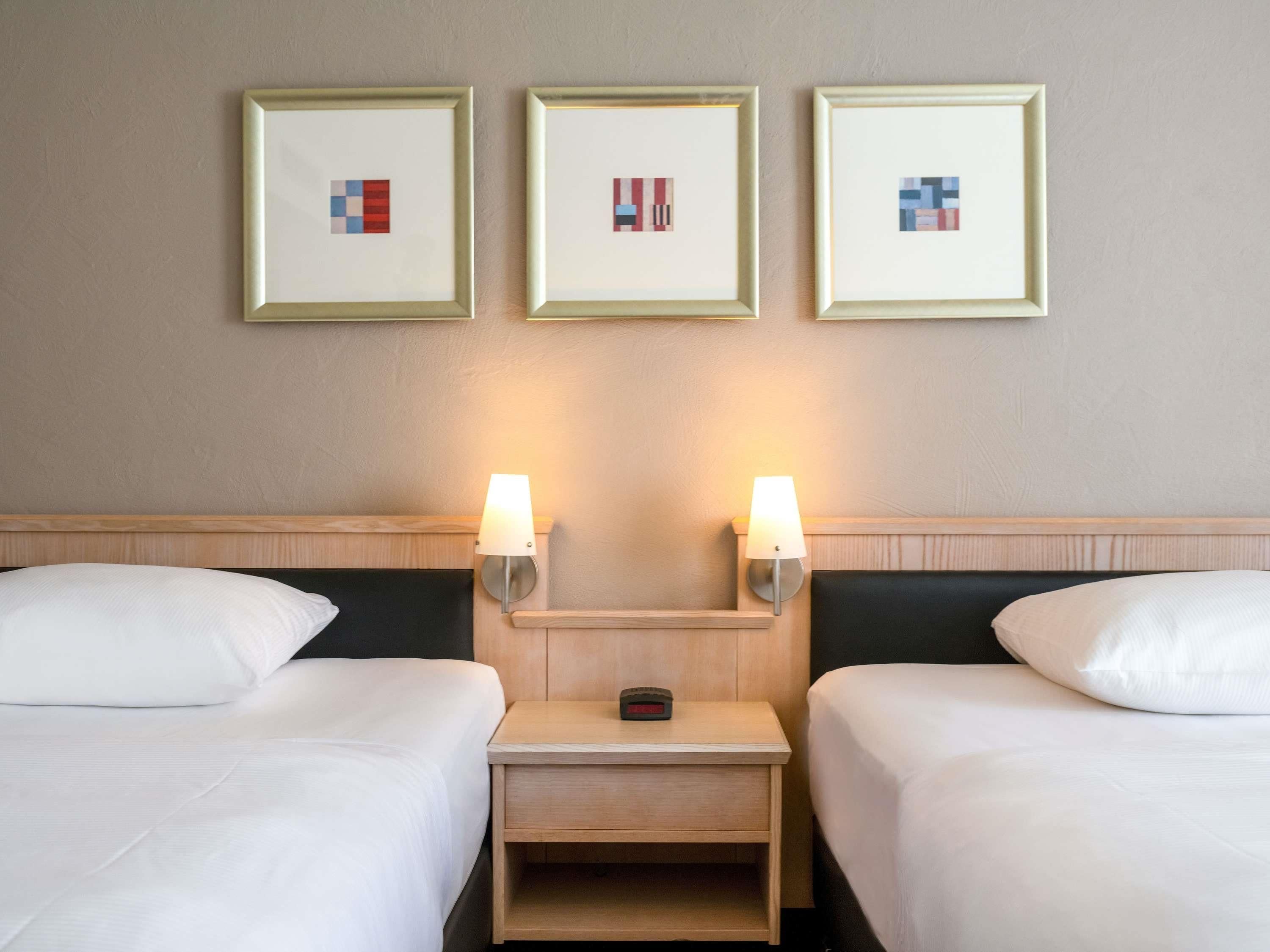 A hotel room with two beds, a nightstand, and three framed pictures on the wall.