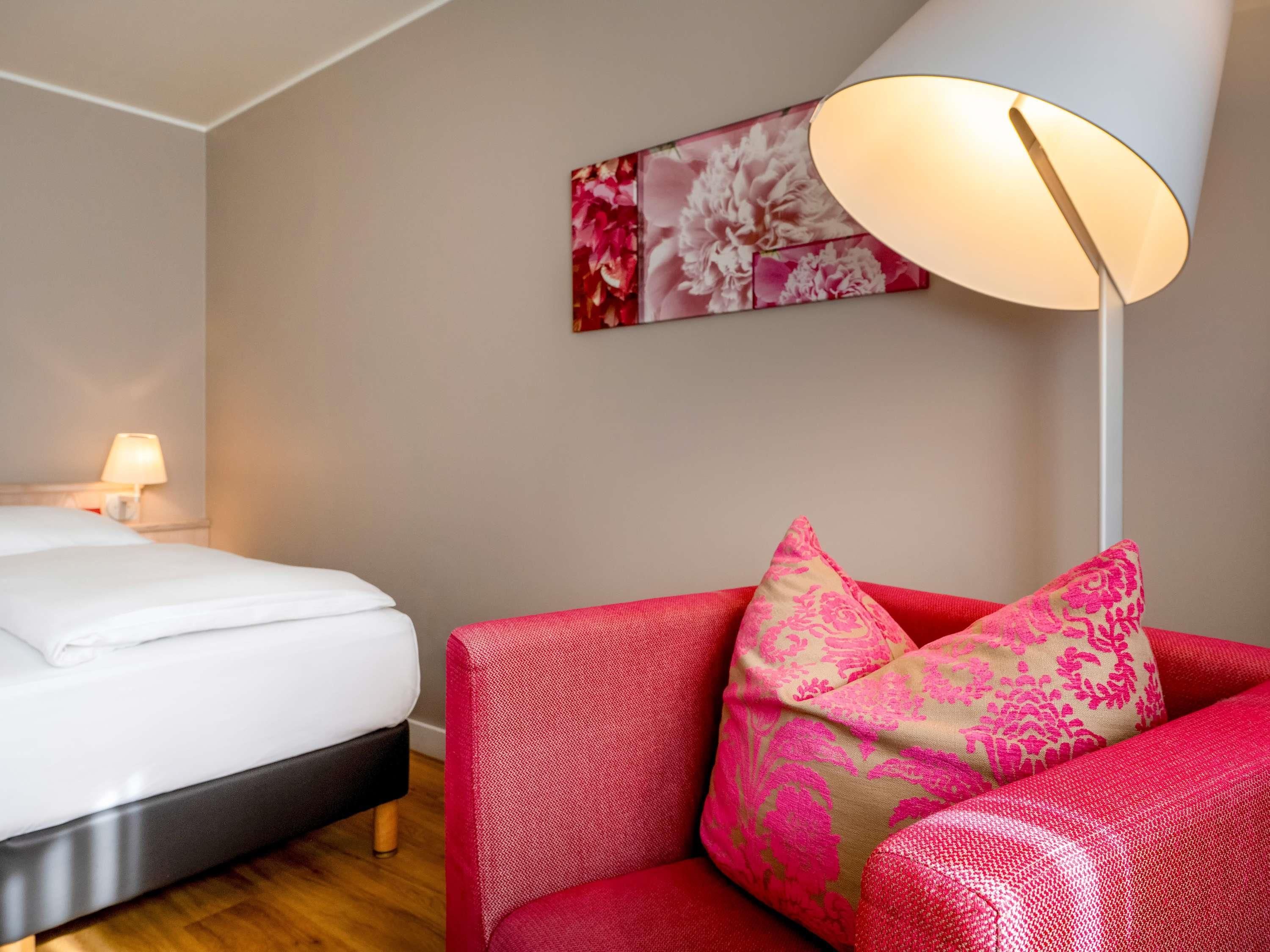Une chambre d’hôtel avec un lit, un canapé rouge orné de coussins à motifs, un lampadaire blanc et une œuvre d’art florale accrochée au mur.