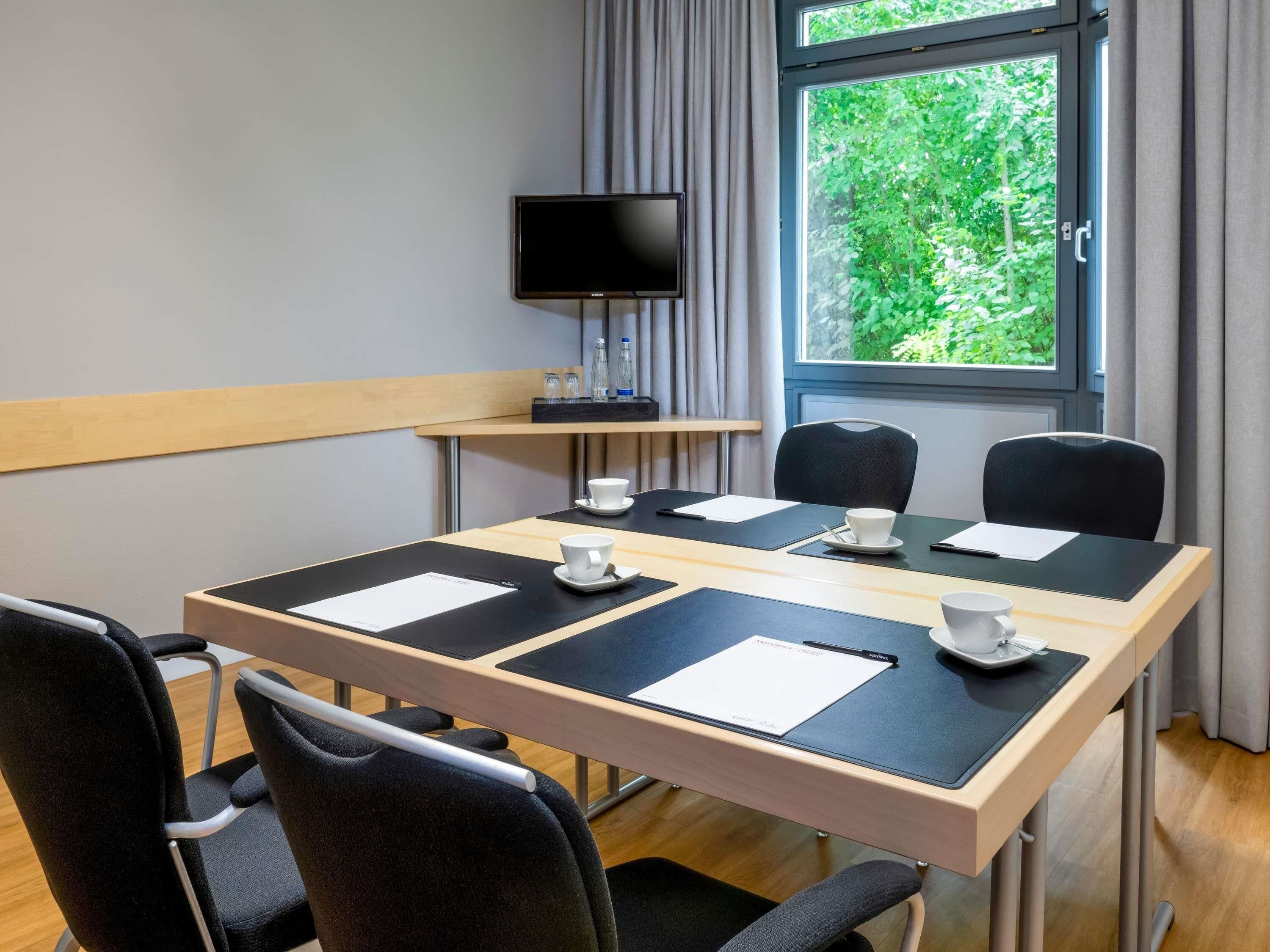 A conference room with a large table, four chairs, a TV, and a window with a view of greenery.