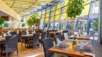 Un restaurante con mesas dentro, ventanales amplios y plantas en macetas.