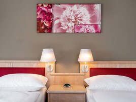 Quarto de hotel com duas camas, uma mesa de cabeceira com relógio digital e luminárias fixadas na parede.
