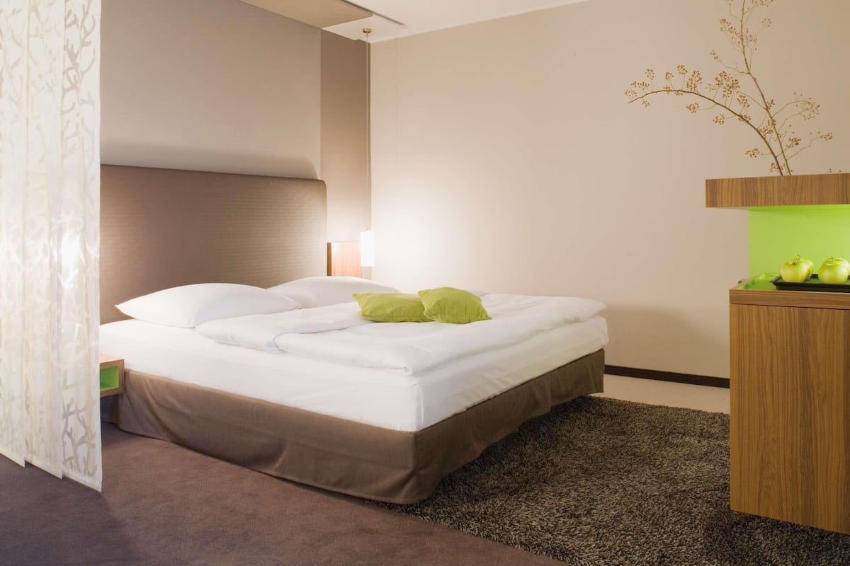 A modern hotel room with a bed, a nightstand, a lamp, and a decorative plant.