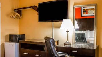 A hotel room with a flat-screen TV mounted on the wall, a desk with a lamp, a chair, a small refrigerator, and a mirror with a red frame.