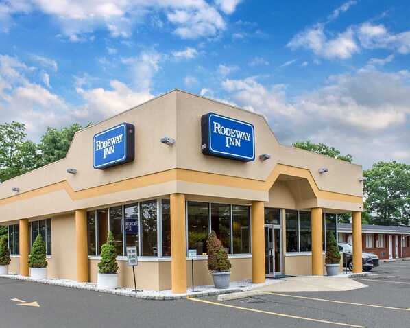 A Rodeway Inn hotel with a beige facade, large windows, and a parking lot. - Rodeway Inn Macarthur Airport (Ronkonkoma)