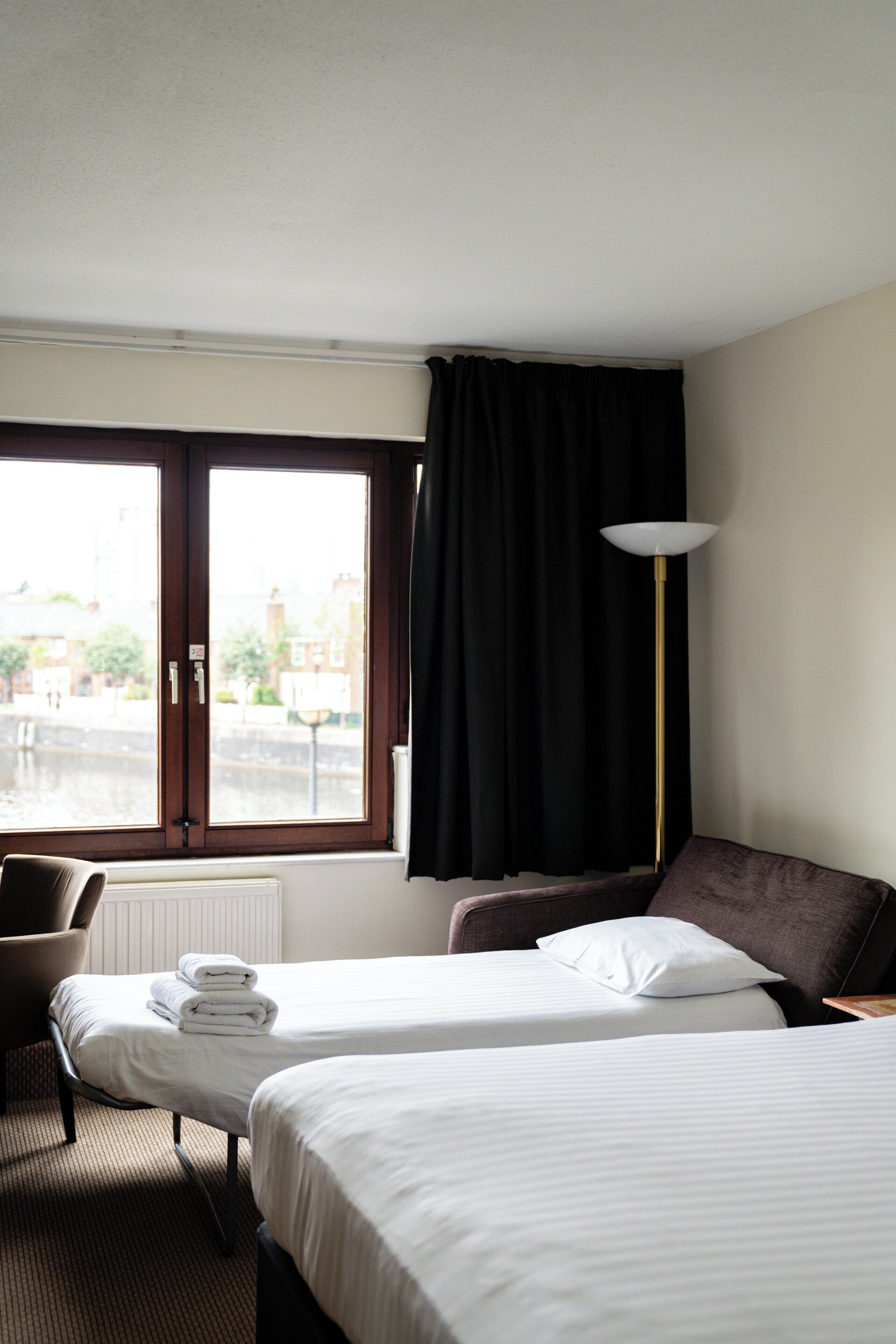 A hotel room with two beds, a chair, a radiator, a window with curtains, and a floor lamp.
