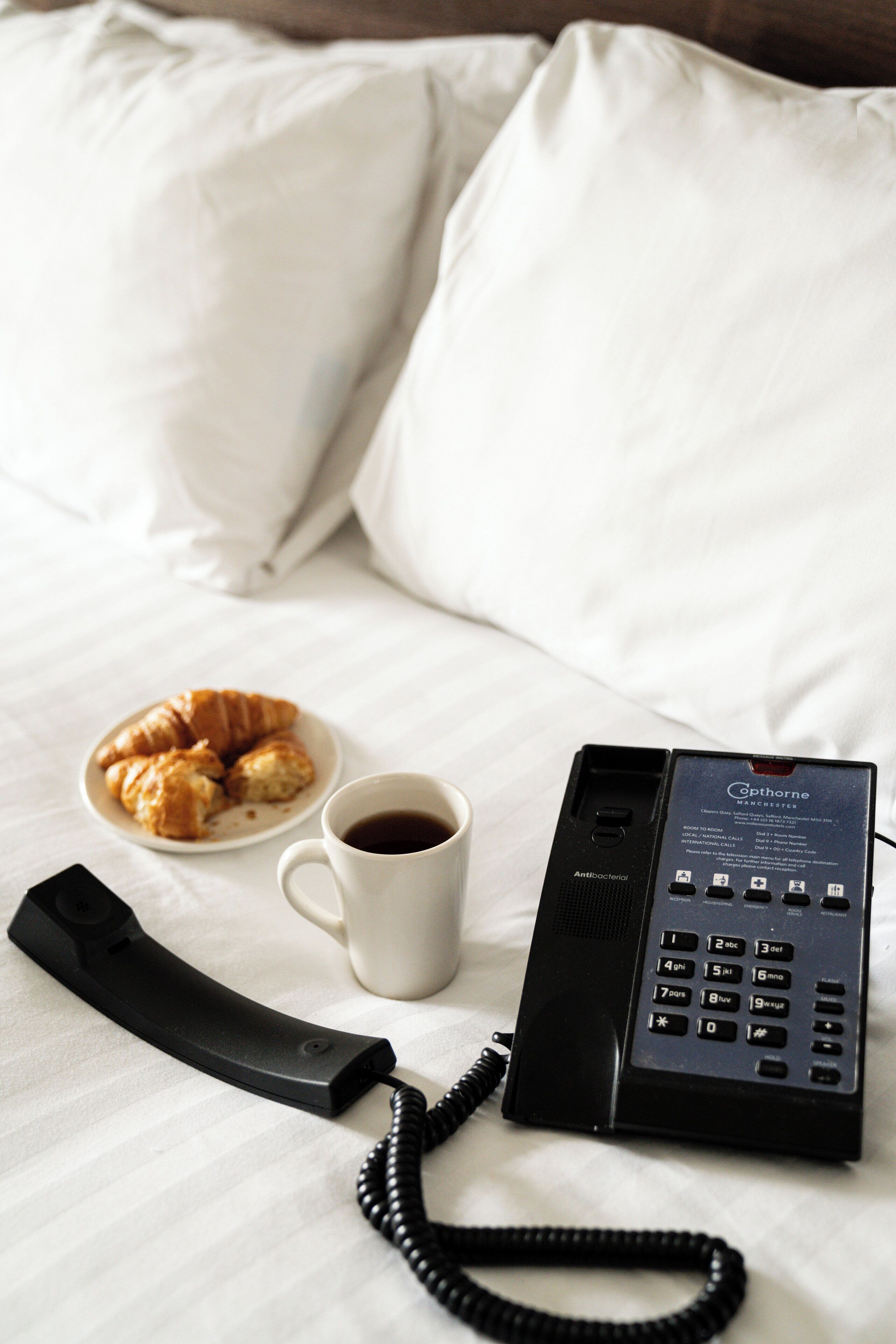Una habitación de hotel con una cama, un plato de croissants, una taza de café y un teléfono.