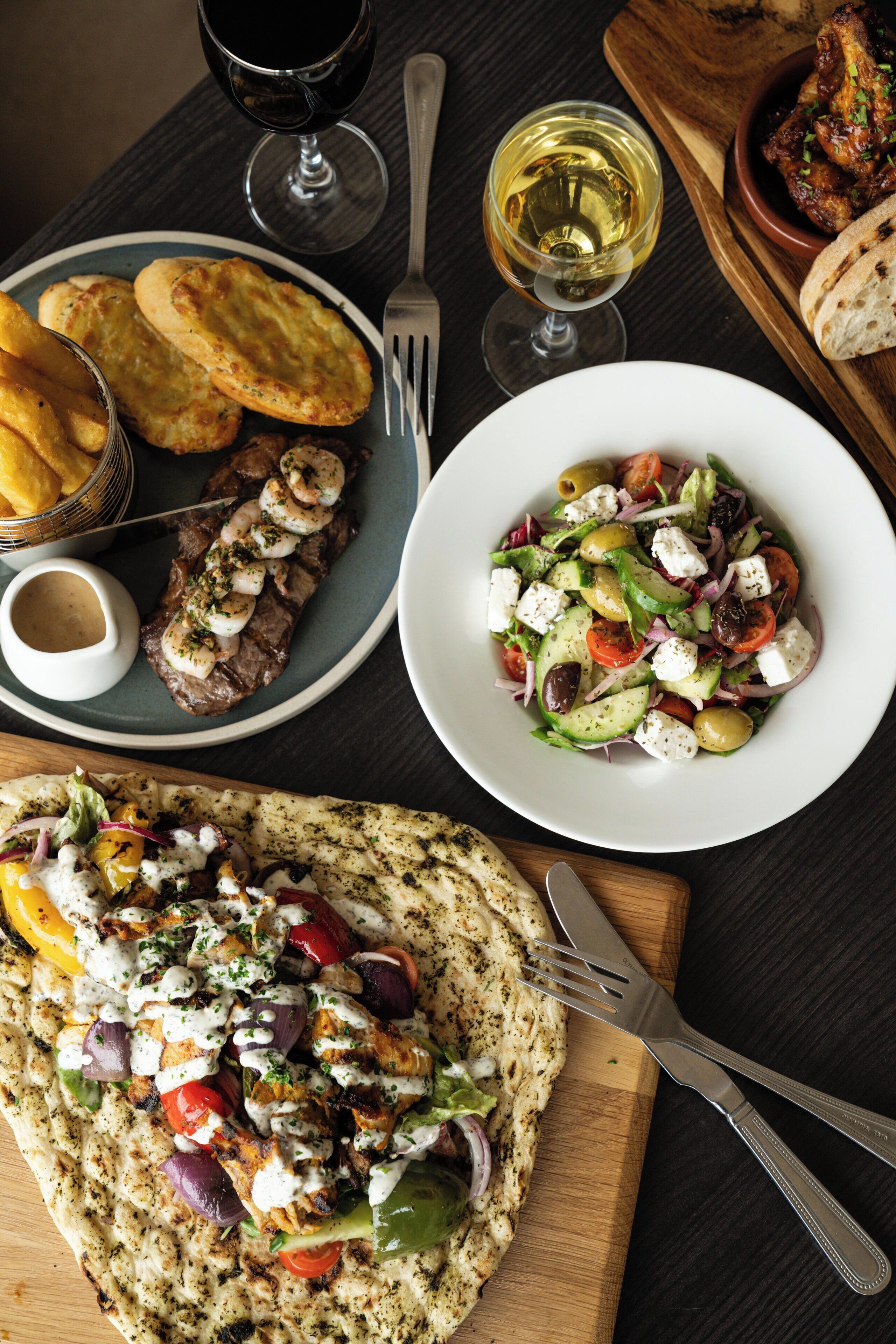 A table with a variety of dishes including grilled meat, a salad, and flatbread, accompanied by glasses of wine.