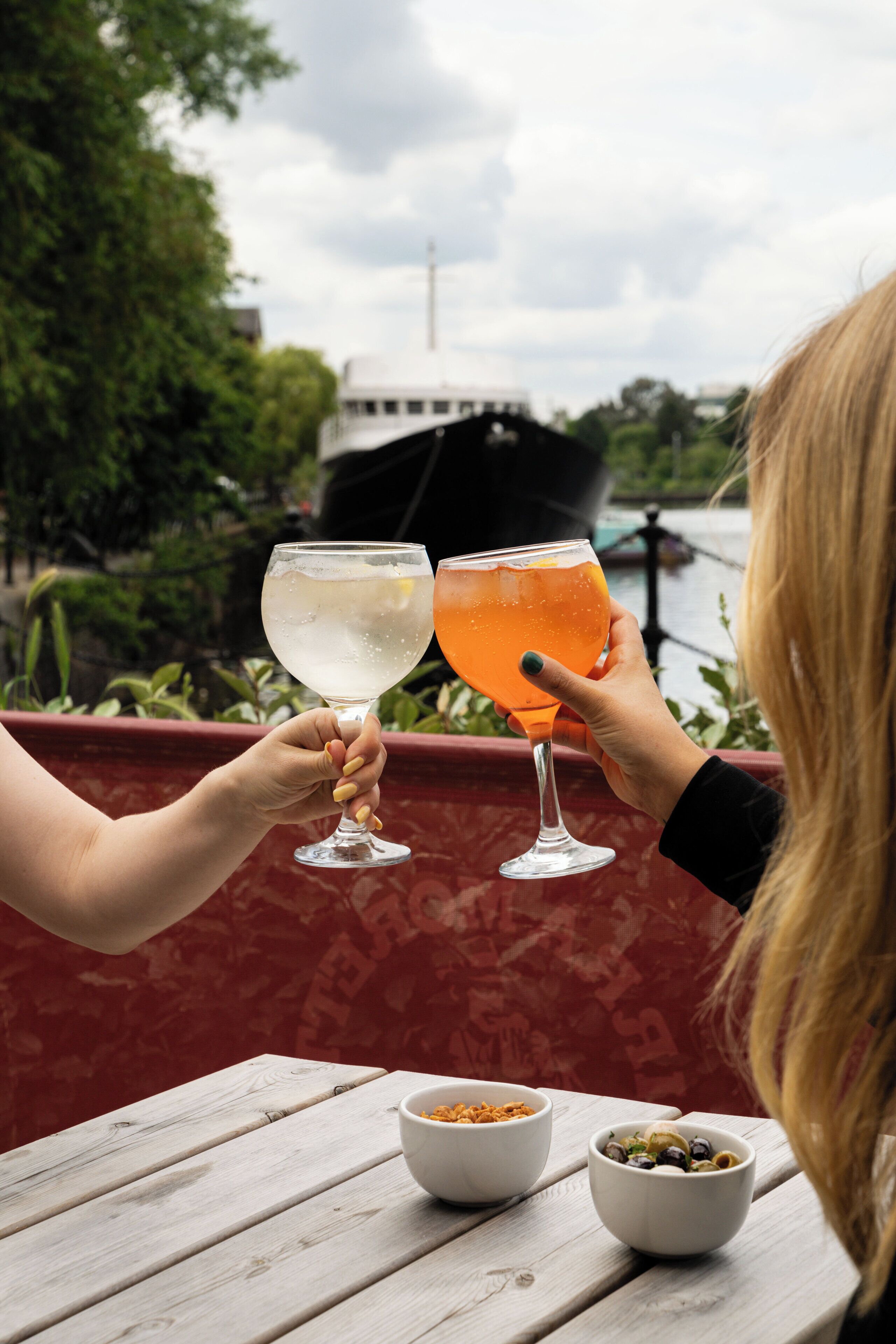 Two people toasting with drinks at a table with bowls of food, overlooking a body of water and greenery.