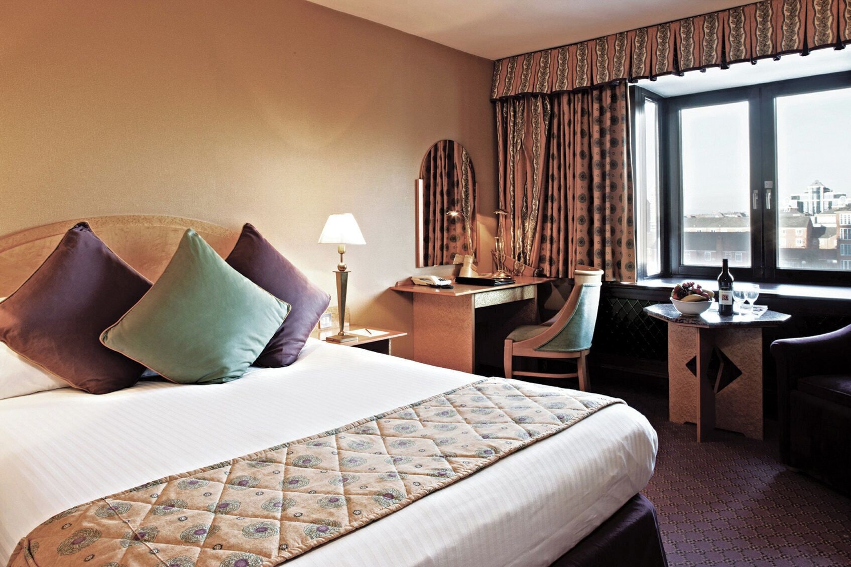 A hotel room with a large bed, a desk, a chair, and a view of the city through the window.