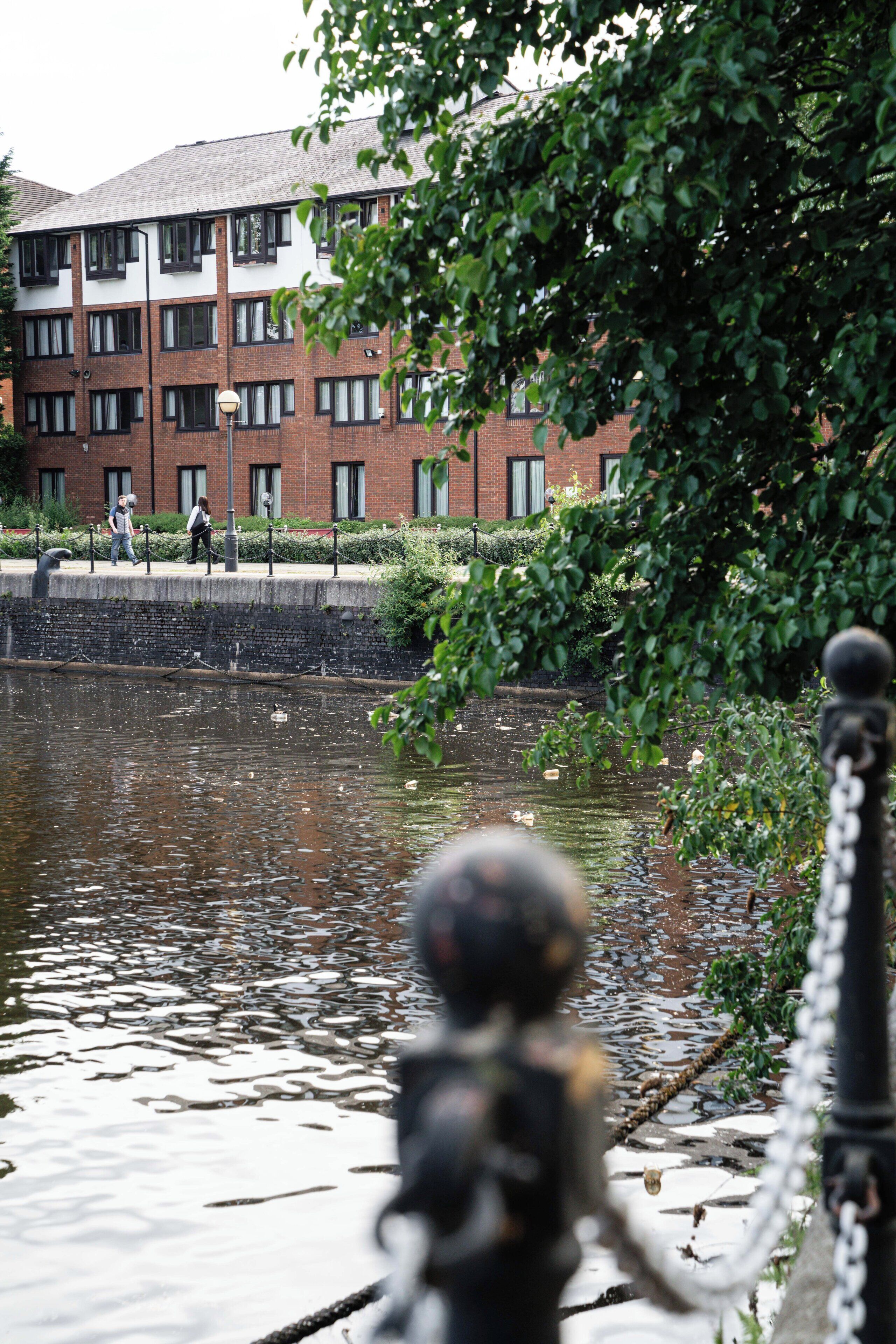 A canal with a statue in the foreground, a brick building in the background, and a pedestrian walkway.