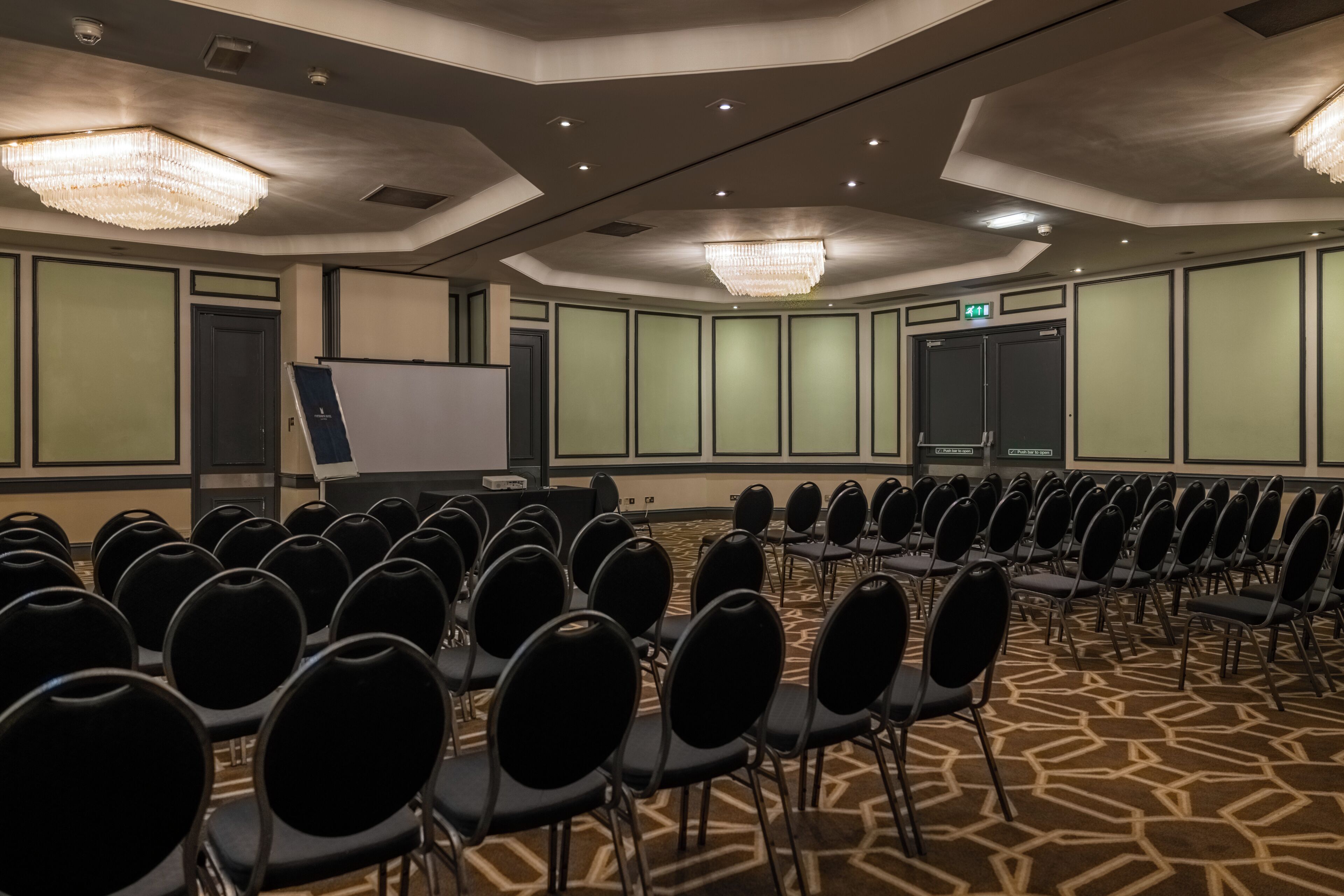Un salón de conferencias con filas de sillas negras, una pizarra y una araña.