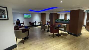 A modern hotel lobby with wooden flooring, several tables and chairs, a flat-screen TV mounted on the wall, and a green plant display.