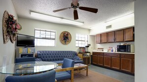 A living room with a glass-top table, blue chairs, a tufted sofa, a flat-screen TV, a ceiling fan, and a kitchen area with wooden cabinets and appliances.