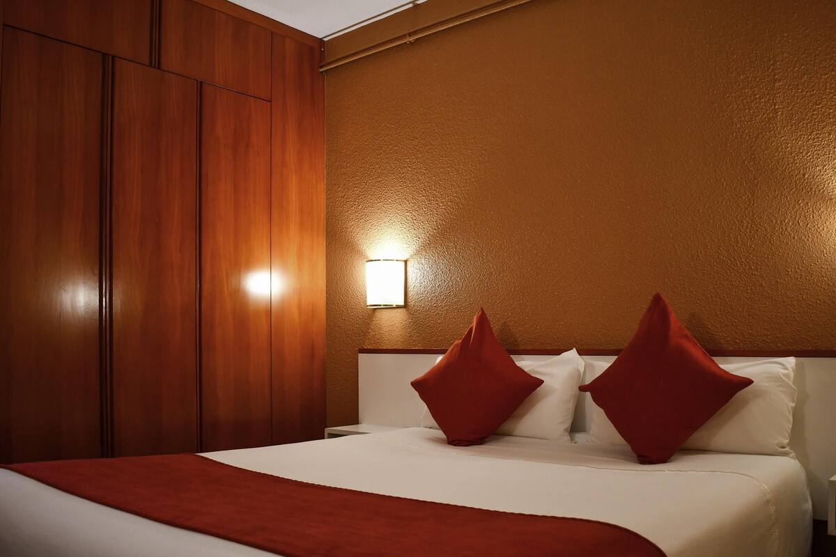A hotel room with a large bed, wooden wardrobe, and a wall-mounted light.