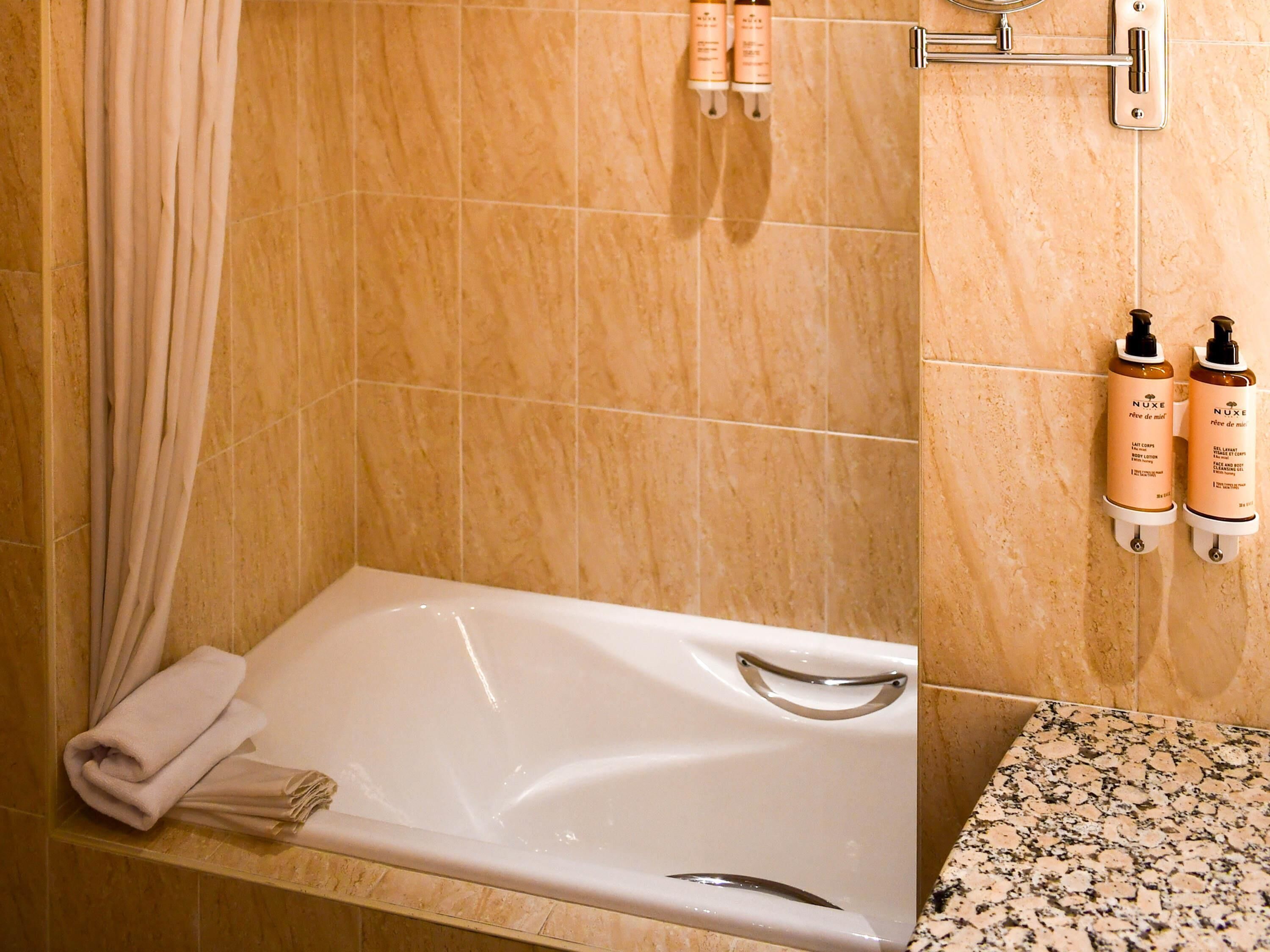 Ein Badezimmer mit Badewanne, einem Handtuchhalter mit Handtüchern und zwei Nuxe Pflegeprodukten an der Wand.