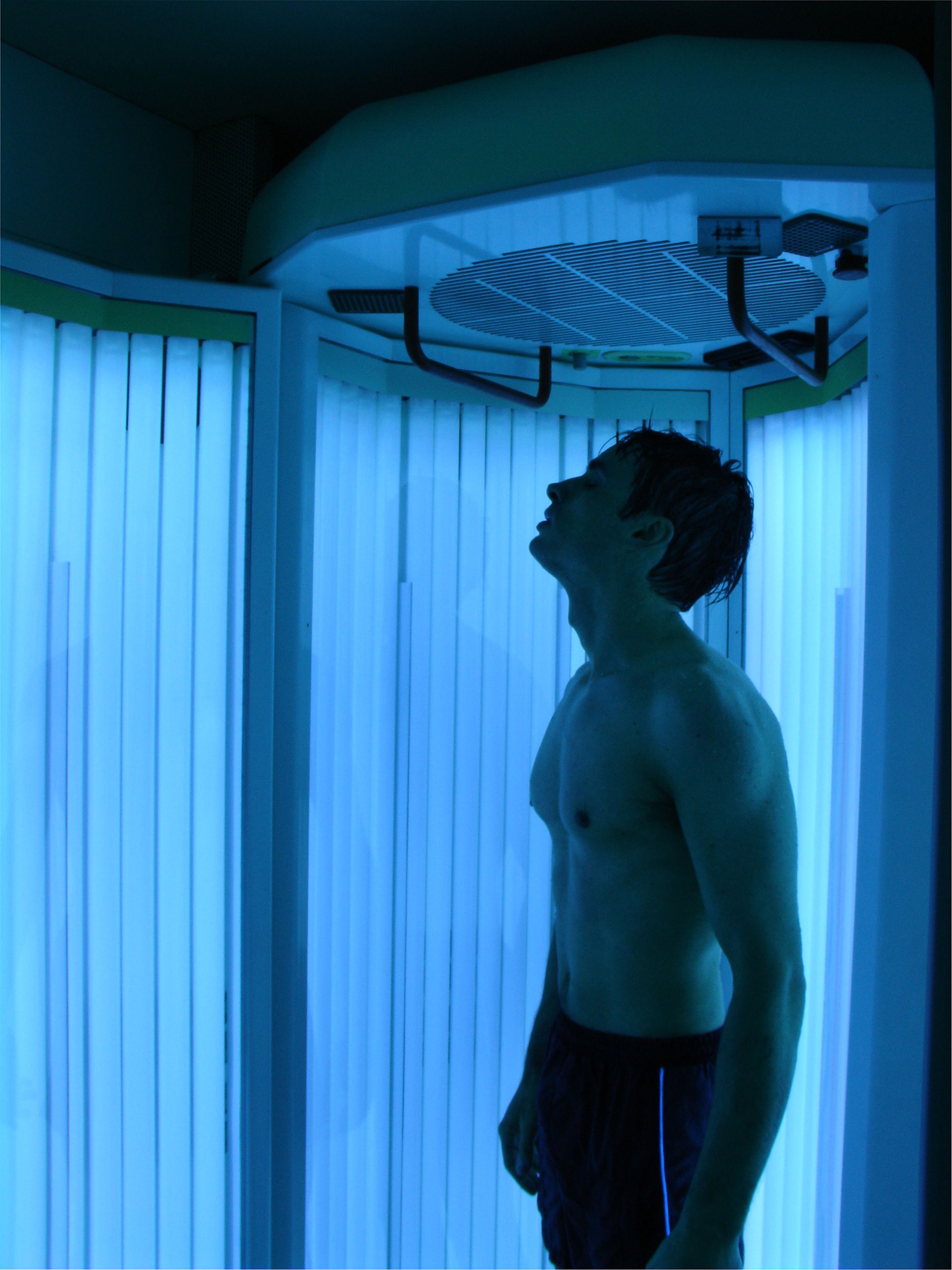 A person standing in a room with a large window and blinds, a ceiling fan, and a light fixture.