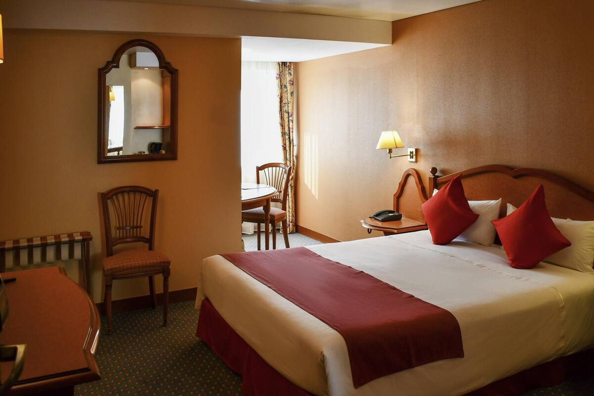 A hotel room with a large bed, a desk, a chair, a mirror, and a lamp.