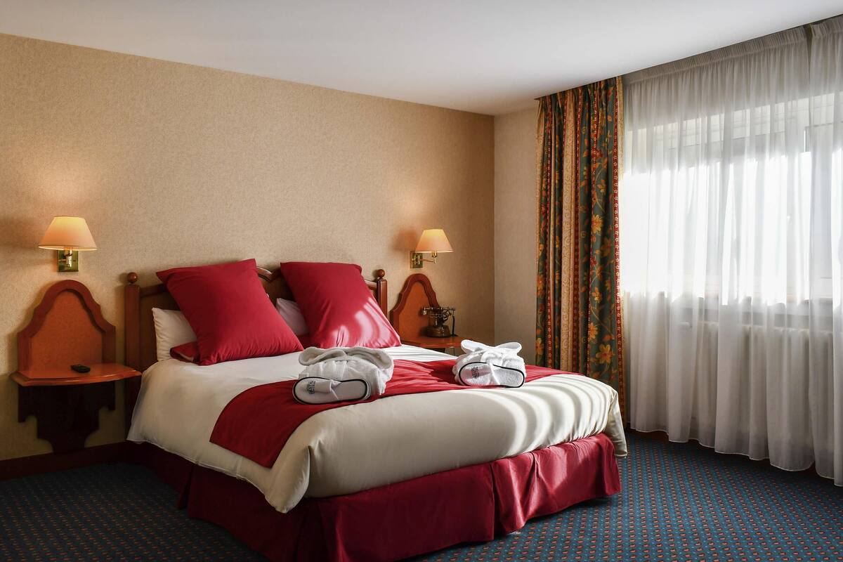 A hotel room with a large bed, red bedding, two bedside lamps, a nightstand, and a window with sheer curtains.