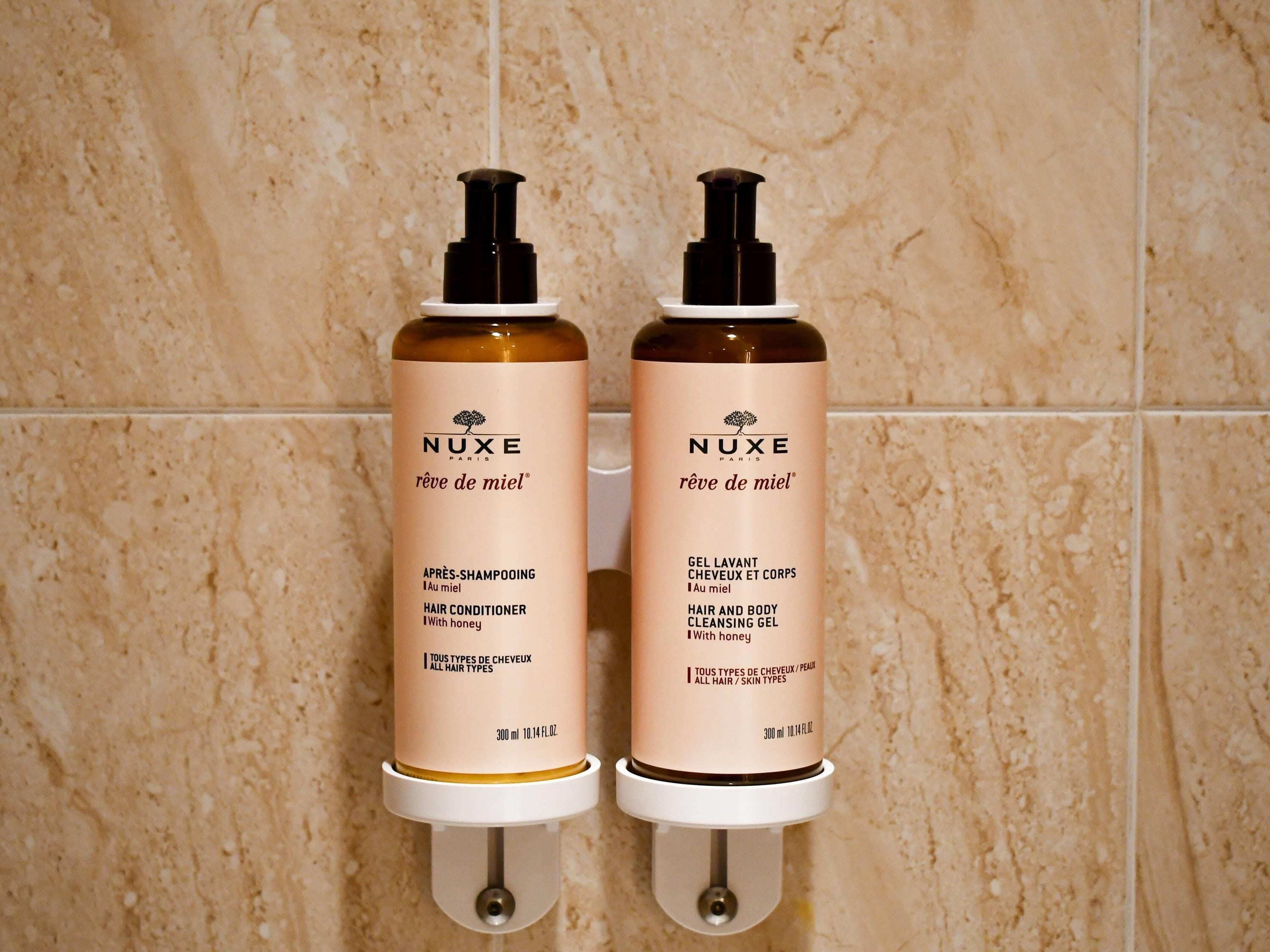 Dois produtos de cuidado capilar Nuxe em um dispensador fixado na parede.