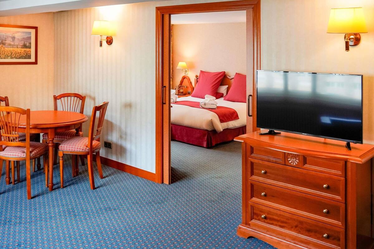 A hotel room with a dining table, chairs, a television, a bed with red bedding, and a painting on the wall.