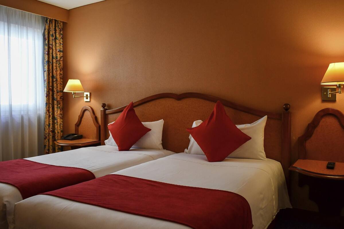 A hotel room with two beds, a nightstand with a lamp, a chair, and a small table.